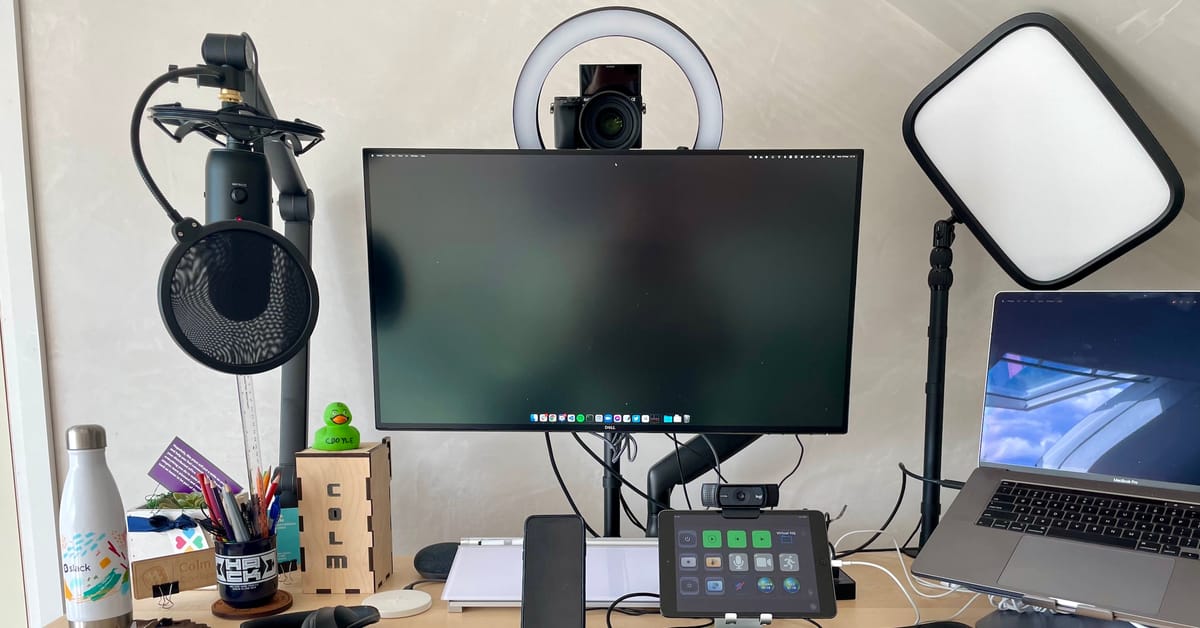 A desk with various items on it, including a microphone, a monitor and a DSLR as a camera.